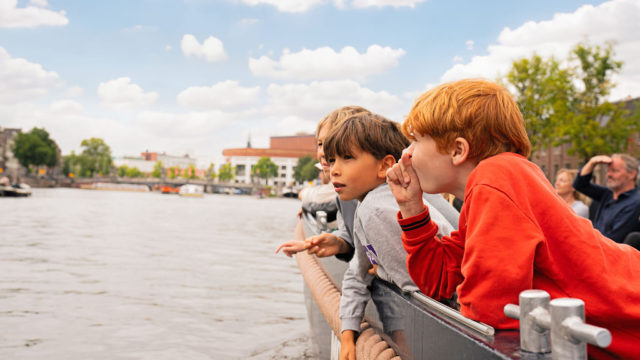 Two boys lean over the edge of a boat, looking out at the calm canal and the trees lining the shore.